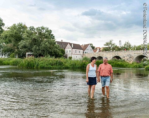 Wanderer in der Wörnitz bei Harburg Ein Paar watet am Wörnitzstrand durchs Wasser. Im Hintergrund sieht man die steinerne Brücke und Häuser von Harburg.