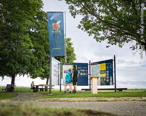 Infostelle bei Kirchheim am Ries Zwei Personen betrachten Informationstafeln im Geopark Ries. Eine weitere Person sitzt auf einer Bank im Hintergrund.