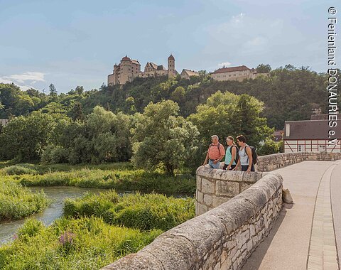 Wandern Harburg Drei Wanderer stehen auf der steinernen Brücke und blicken auf die Wörnitz. Links oben thront über dem Wald die Harburg.
