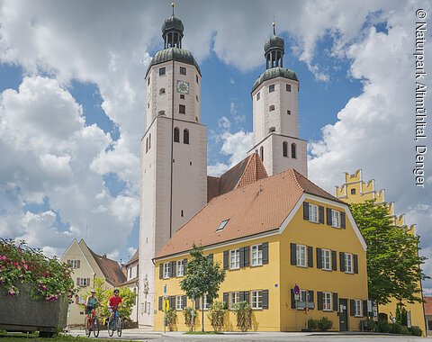 Radfahrer in Wemding Zwei Radfahrer vor einem gelben Gebäude und einer Kirche mit zwei Türmen und blauen Himmel im Hintergrund.
