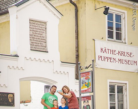 Ferienland Donau-Ries Shooting 2021 Eine Familie macht ein Selfie vor dem Käthe-Kruse-Puppen-Museum in einem gelben Gebäude mit Schildern.