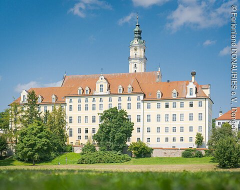 Heilig Kreuz Garten in der Altstadt von Donauwörth Großes, mehrstöckiges Gebäude mit orangefarbenem Dach und Turm, umgeben von Bäumen und Wiese, unter blauem Himmel.