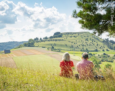 Der Ipf vom Blasienberg aus Zwei Personen sitzen auf einer Wiese und blicken auf eine hügelige Landschaft mit Feldern und Bäumen.
